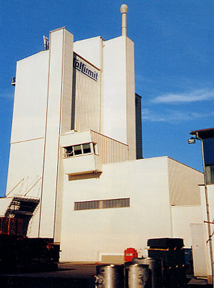 Hohes, helles Industriegebäude mit mehreren vertikalen Baukörpern und einem kleinen, vorragenden Kontrollraum; auf der Fassade ist der Firmenname sichtbar, unter einem klaren blauen Himmel.
