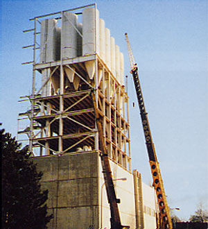 Großer Industriebau mit einem hohen Stahlgerüst und mehreren vertikalen Silos; daneben steht ein Mobilkran, der an der Konstruktion arbeitet.