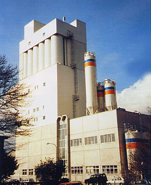 Das Bild zeigt eine industrielle Produktionsanlage mit einem hohen, mehrstöckigen Gebäude, das mehrere große silberne silos an seiner Seite aufweist. Diese Silos sind farblich markiert, um sich visuell abzuheben. Die Fassade des Gebäudes weist große Fensterflächen auf, die vermutlich Büros oder Verwaltungsräume beherbergen. Auf der unteren Ebene sind verschiedene Fahrzeuge zu sehen, die vor der Anlage geparkt sind.