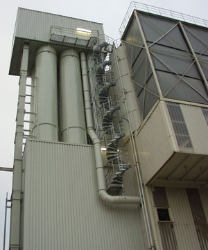 Außenansicht einer Industrieanlage mit mehreren vertikalen Silorohren und einer seitlich verlaufenden Metallwendeltreppe, angebracht an einer hellen Stahlkonstruktion.