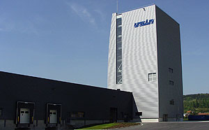 Modernes Industriegebäude mit niedrigem Hallentrakt und einem hohen, silbergrauen Turm, auf dem ein Firmenlogo steht; im Vordergrund befinden sich Laderampen unter klarem Himmel.