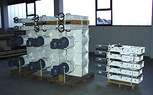 Industriebauteile für Absperrsysteme in einer Werkhalle: links mehrere große, weiß lackierte Absperrschieber mit Antrieben, rechts gestapelte flache Metallmodule auf Paletten.