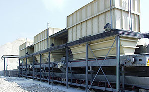 Lange Bandanlage mit mehreren großen Beschickungsbehältern auf einem erhöhten Stahlgerüst, umgeben von hellem Schüttgut in einer offenen Industrieumgebung.