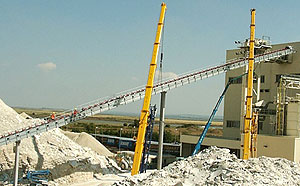 Schräg verlaufendes Förderband zwischen zwei gelben Stützgerüsten, installiert in einem weitläufigen Außenbereich mit Schüttguthalden und Gebäuden im Hintergrund.