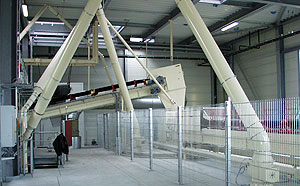 Innenansicht einer Bandanlage mit mehreren hellen, schräg verlaufenden Förderrohren und Gestellen, abgesichert durch ein umlaufendes Metallgitter.