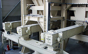 Nahaufnahme mehrerer parallel angeordneter Trogkettenförderer aus hellem Metall, montiert unter einer großen Stahlkonstruktion.