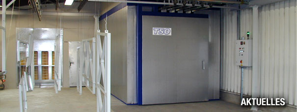 Innenansicht einer Anlage zur Pulverbeschichtung: links hängen vorbereitete Metallteile an einer Laufbahnschiene, rechts steht eine große, geschlossene Pulverbeschichtungskabine mit Bedienfeld.